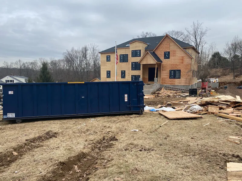 Blue 30 yard dumpster on a construction site in front of a USA flag pole and house under construction
